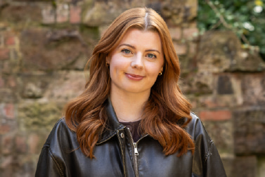 ead and shoulders outdoor portrait of a white woman with red hair, smiling and wearing a brown jacket, positioned against a brick wall in natural light. 