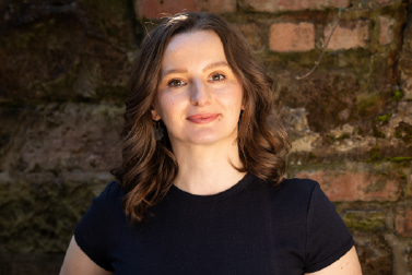 Head and shoulders outdoor portrait of a white woman with brown hair, smiling and wearing a black T-shirt, positioned against a brick wall in natural light.