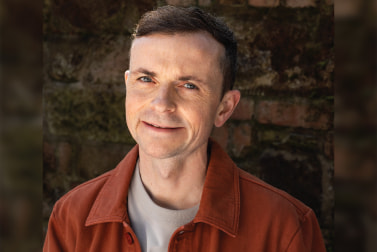 Head-and-shoulders outdoor portrait of a white man with short dark hair, smiling and wearing a rust-coloured jacket over a light shirt, positioned against a textured brick wall in natural light.