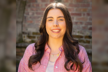 Portrait of a white woman with brown hair, smiling and wearing a stripy, red and white shirt over a t-shirt. Positioned against a brick wall with natural light.