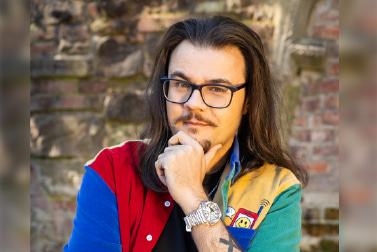 Portrait of a mixed raced man with long dark brown hair, smiling to camera, wearing a yellow and red shirt with blue and green rolled up sleeves, positioned against a brick wall in natural light.
