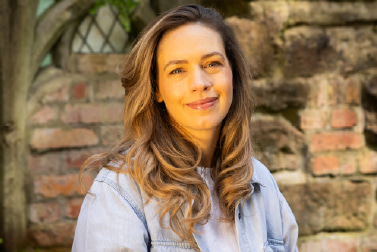 Head and shoulders outdoor portrait of a white woman with light brown/ blond hair, smiling and wearing a white dress and a light blue denim shirt, positioned against a brick wall in natural light.