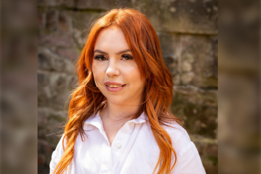 An image of white women with long wavey gingery orange hair, smiling to camera wearing a white blouse stood in front of a brick wall with natural sunlight. 