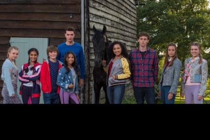The main Free Rein characters are standing in a line, with a horse in their middle. Fields and a barn inthe background.