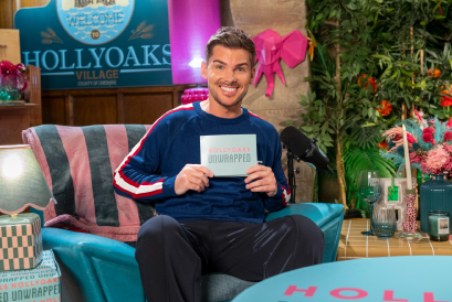 Kieron Richardson sits in the Hollyoaks Unwrapped set with cue cards in his hand, smiling to the camera.