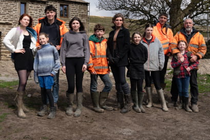 Picture of Amanda Owen and her family standing in a field outside. the family made up of Amanda, partner Clive and their 9 children, stand in a line, looking towards the camera there are fields and hills in the background.