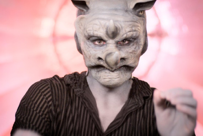 A person wearing a lifelike, rhinoceros mask looks to the camera wearing a black and white stripe shirt